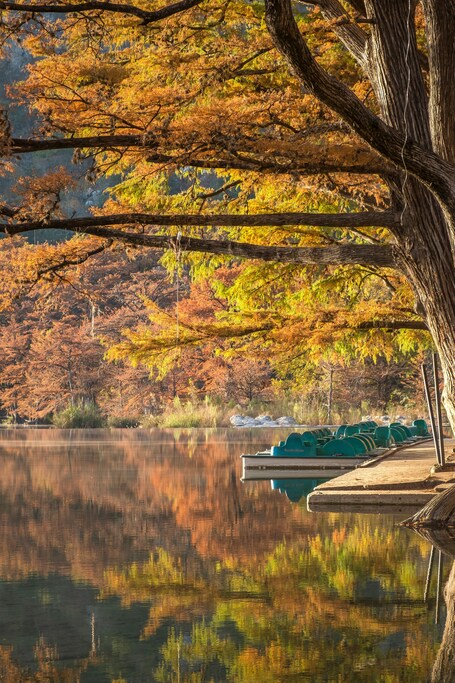 A serene lake with autumn foliage, a dock with boats, and a calm reflection of the trees.