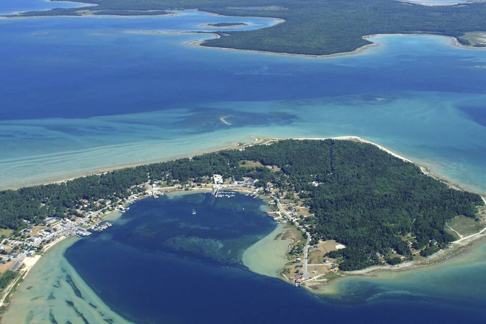 An aerial view of a coastal area with a central peninsula surrounded by water, featuring a dense forest, a marina, and residential buildings.