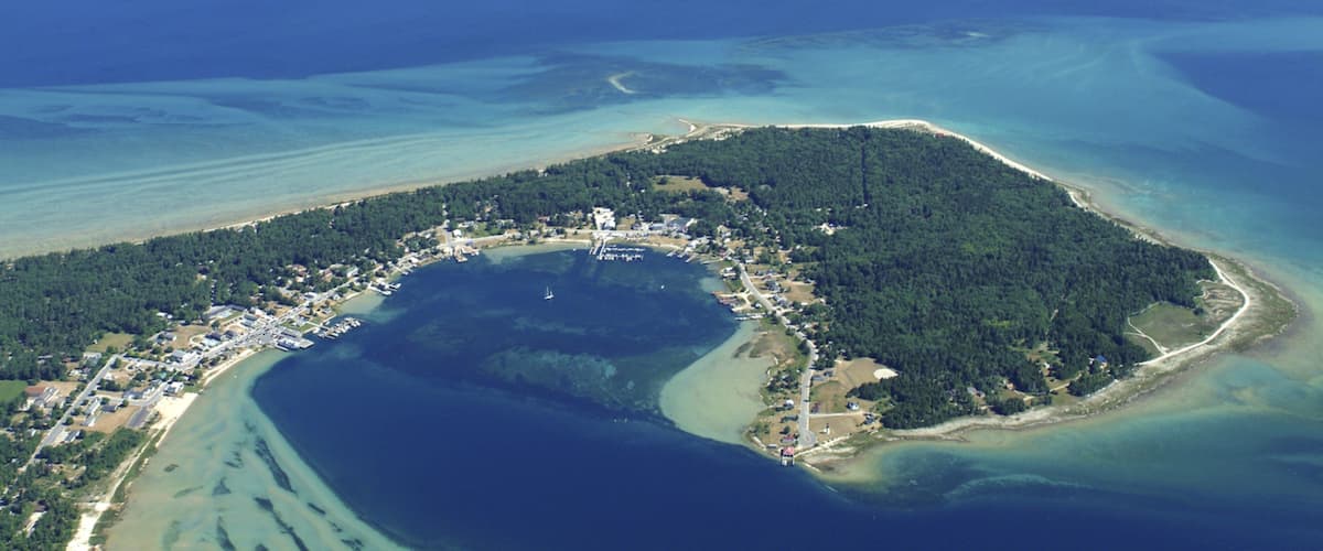 An aerial view of a coastal area with a central peninsula surrounded by water, featuring a dense forest, a marina, and residential buildings.