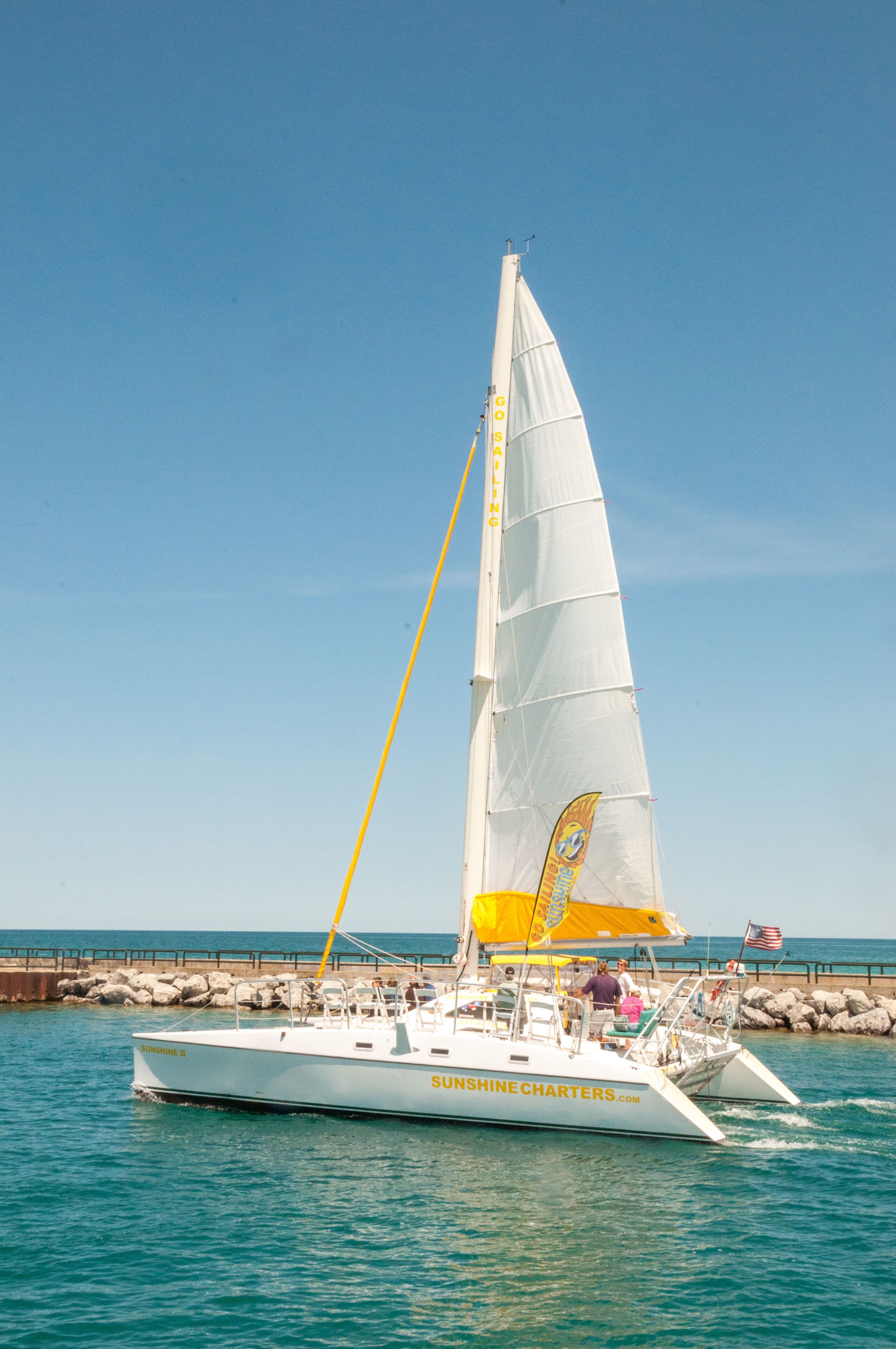 Un velero llamado 'Sunshine Charters' con vela amarilla y blanca, amarrado en un muelle con un cielo azul despejado al fondo.