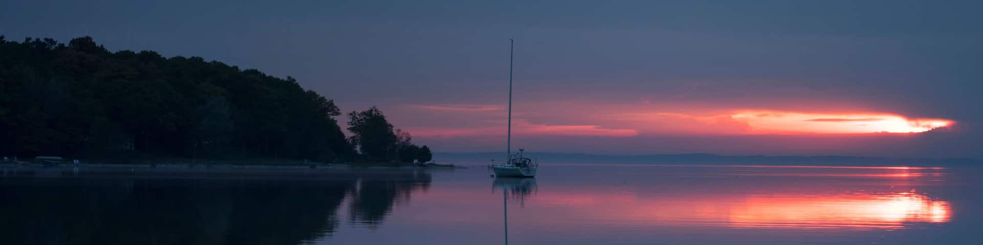 A calm lake at sunset with a sailboat anchored in the center, surrounded by a forested shoreline.