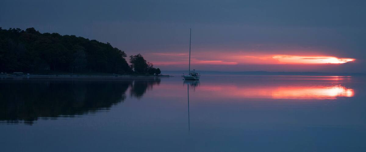 A calm lake at sunset with a sailboat anchored in the center, surrounded by a forested shoreline.