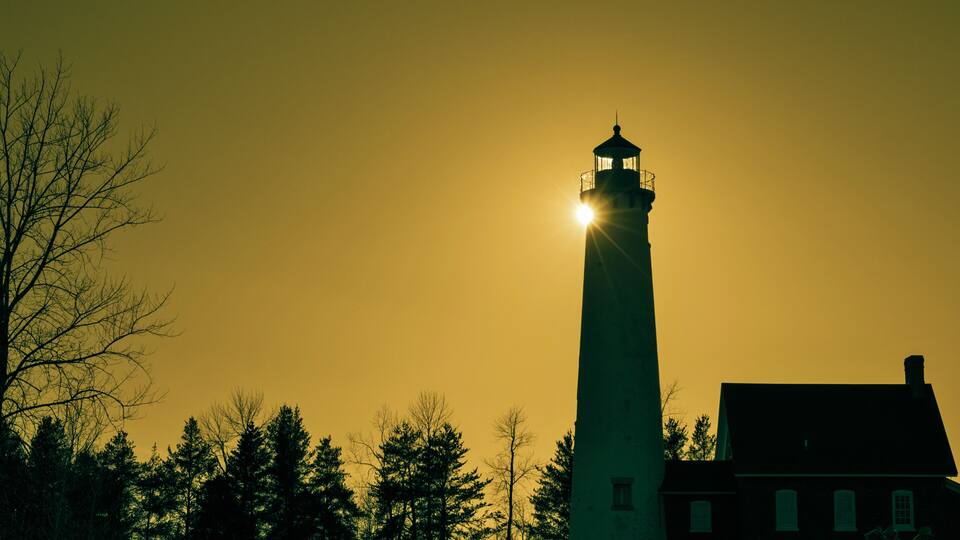A lighthouse with a bright light at its top, surrounded by trees and a building.