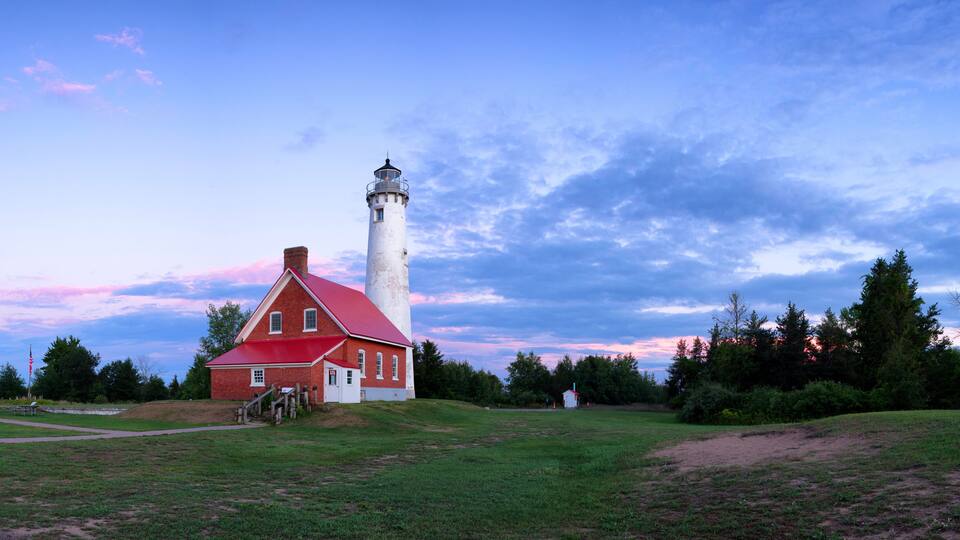 A red brick lighthouse with a white tower, surrounded by a grassy field and trees under a twilight sky.