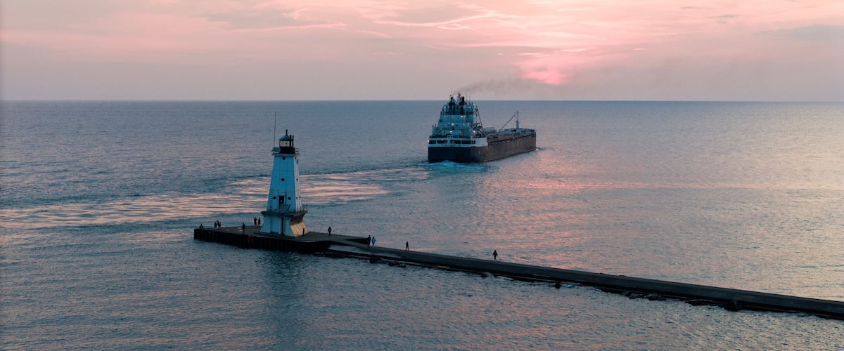 A lighthouse and a cargo ship in the ocean at sunset.