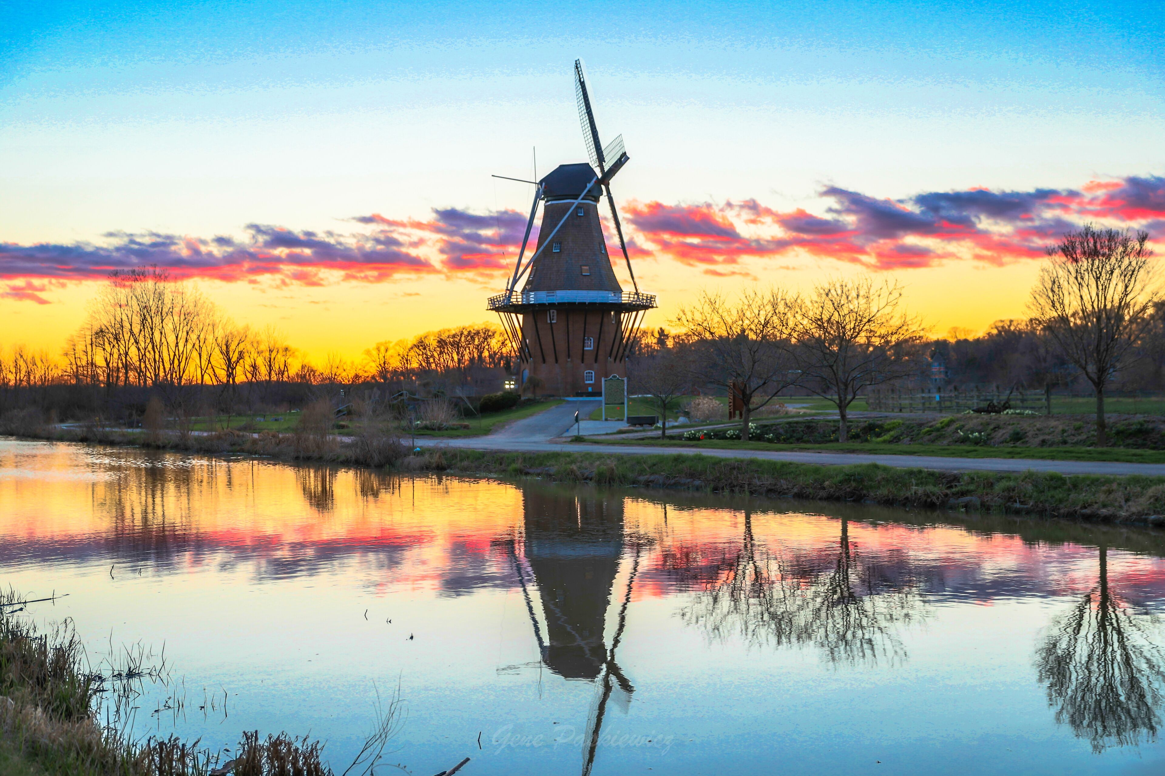 A windmill by a calm lake at sunset with a colorful sky.