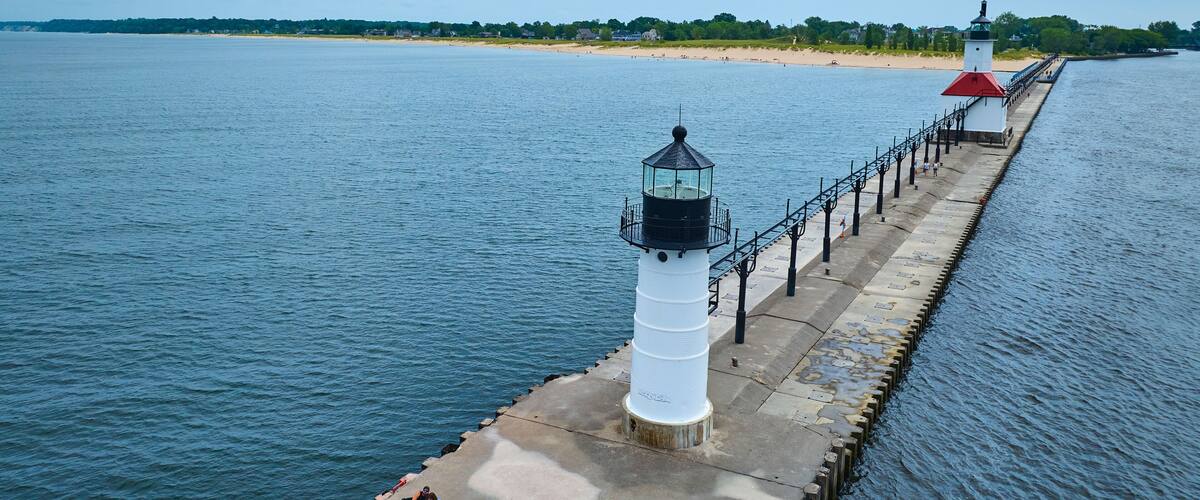 A lighthouse and pier extending into a calm body of water, with a beach and trees in the background.