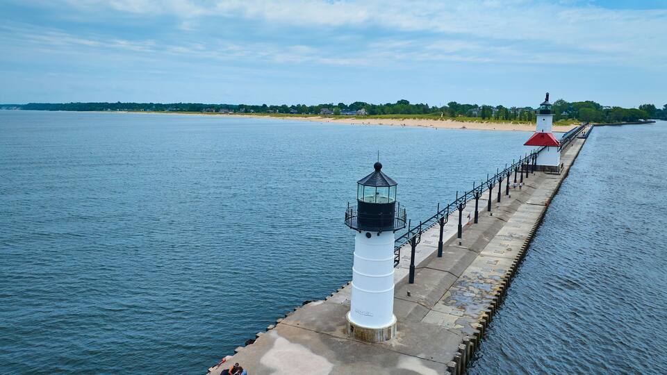 A lighthouse and pier extending into a calm body of water, with a beach and trees in the background.