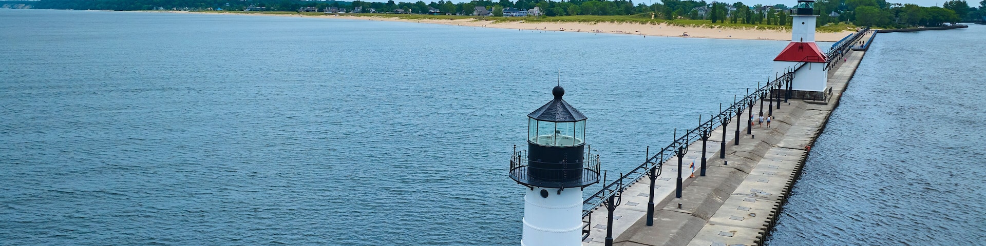 Un faro y un muelle se adentran en aguas tranquilas, con una playa y árboles al fondo.
