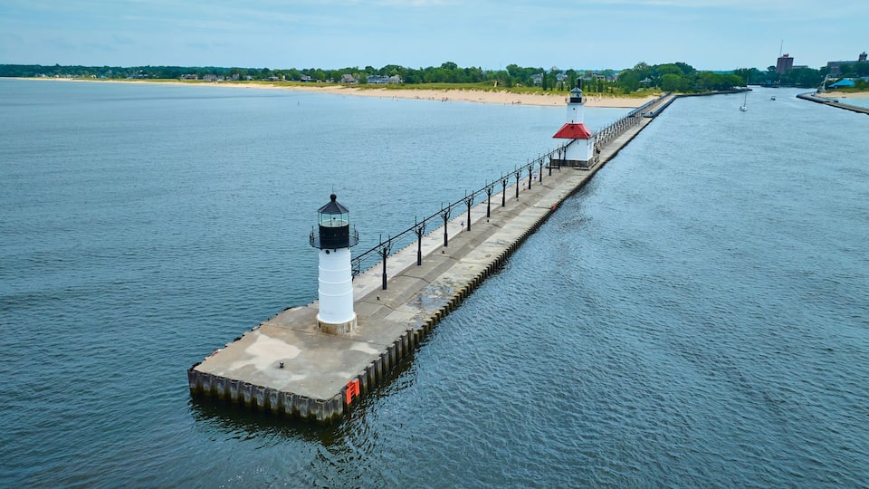 A lighthouse on a pier extending into a calm body of water, with a beach and buildings in the background.