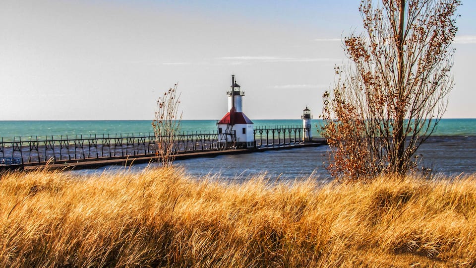 A coastal lighthouse with a red roof, a pier with a black railing, and golden grasses in the foreground.