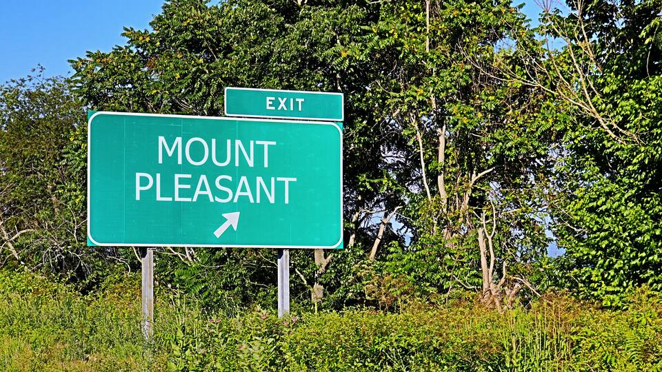 A green road sign with 'MOUNT PLEASANT' and an arrow pointing right, surrounded by lush greenery.
