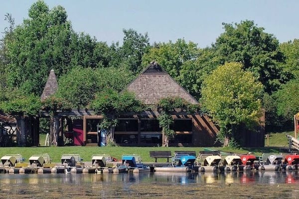A lakeside area with a thatched-roof building, boats, and lush greenery.