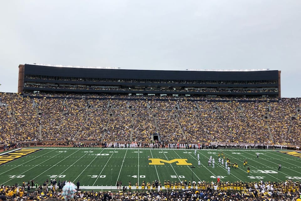 A football stadium filled with spectators, featuring a large field marked with yard lines and a prominent team logo.