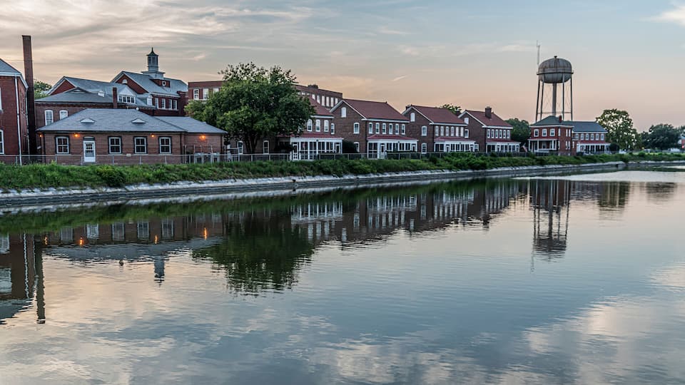 A calm river reflecting buildings and a water tower.