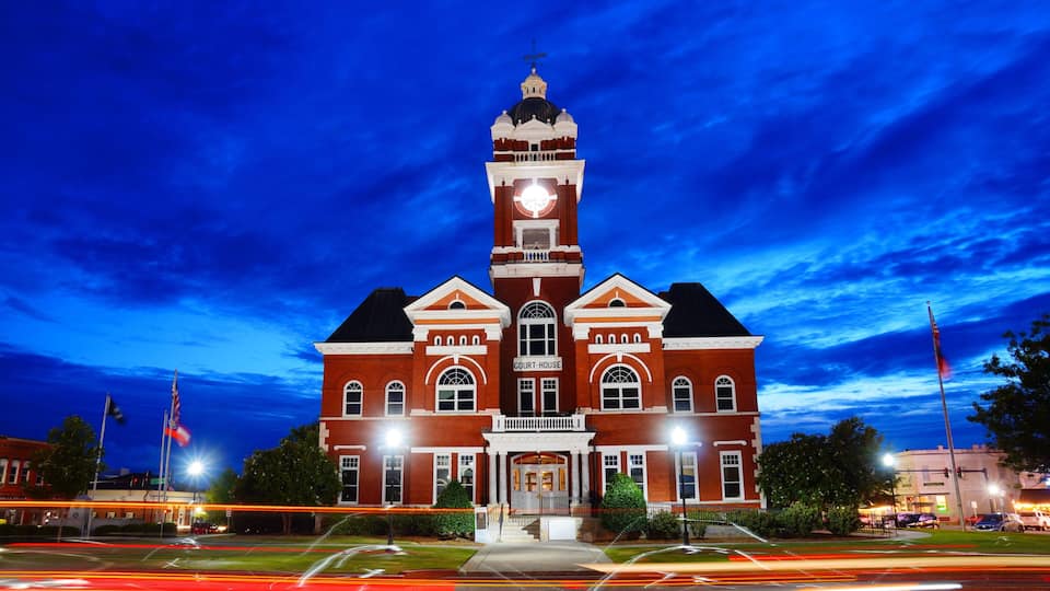 A large red brick courthouse with a clock tower, surrounded by a well-lit street and trees.