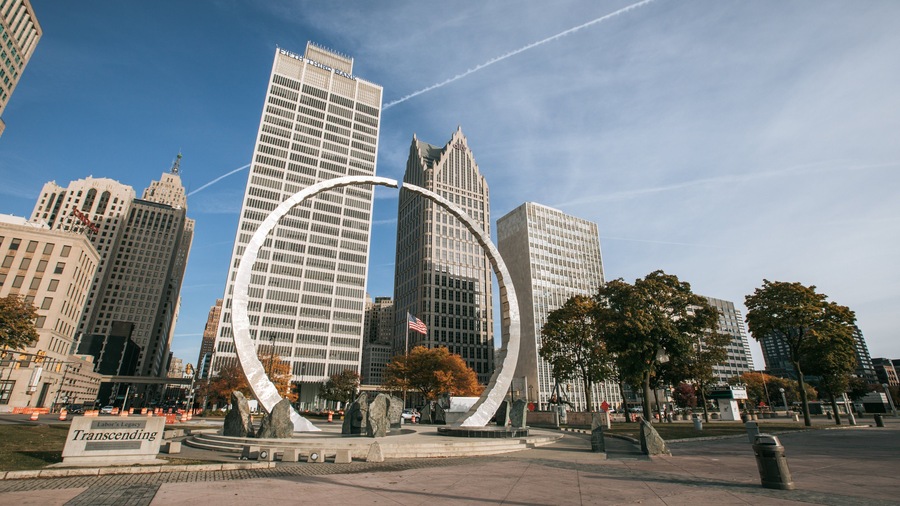 A cityscape with tall buildings, a large circular sculpture, and an American flag.