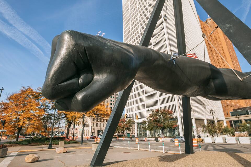 A large metallic fist sculpture in an urban setting with tall buildings and autumn foliage.