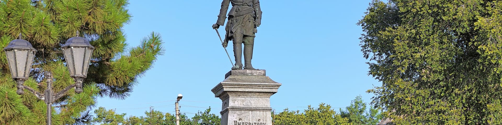 A statue on a pedestal with inscriptions, surrounded by a chain-link fence, trees, and buildings in the background.