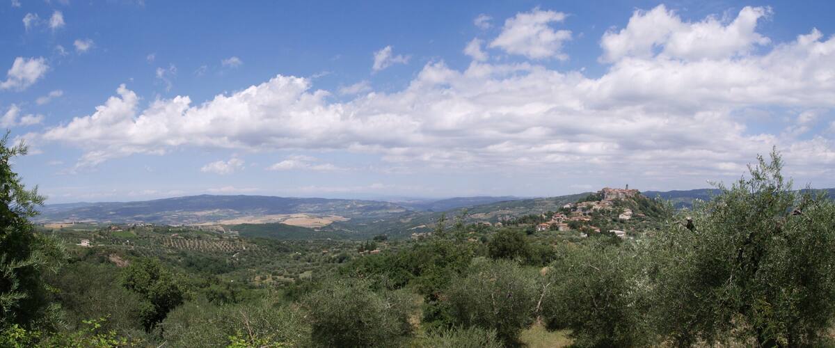 A mountainous landscape with olive groves, a historic castle on a hill, and a clear blue sky with scattered clouds.