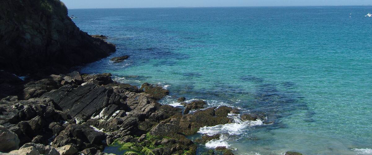 A coastal landscape with clear blue waters, rocky shores, and a grassy cliff with trees.