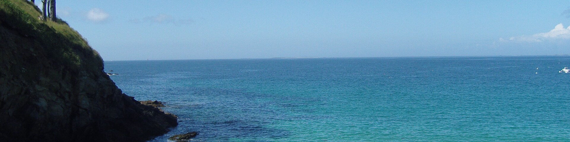 A coastal landscape with clear blue waters, rocky shores, and a grassy cliff with trees.