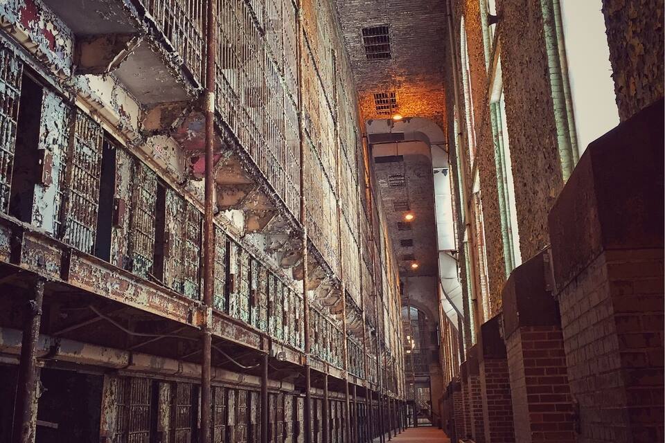 A narrow, dimly lit corridor with high ceilings and metal railings, lined with brick walls and a concrete floor.