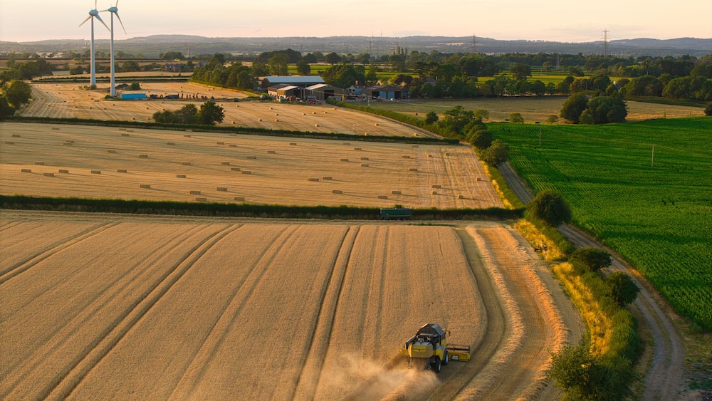 Une moissonneuse-batteuse travaille dans un champ, avec des éoliennes et un paysage rural en arrière-plan.