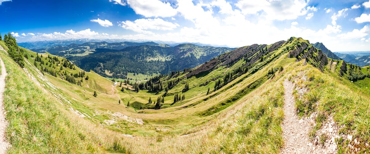 A mountain landscape with a dirt path, green hills, and distant mountains under a blue sky.