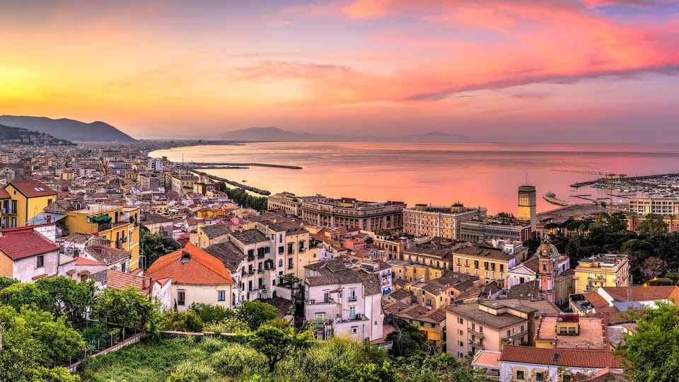 A coastal cityscape with a sunset over the sea, featuring historic buildings and a harbor.