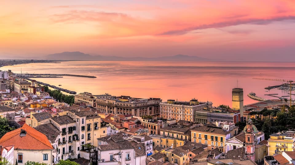 A coastal cityscape at sunset with a view of the sea, buildings, and mountains in the background.