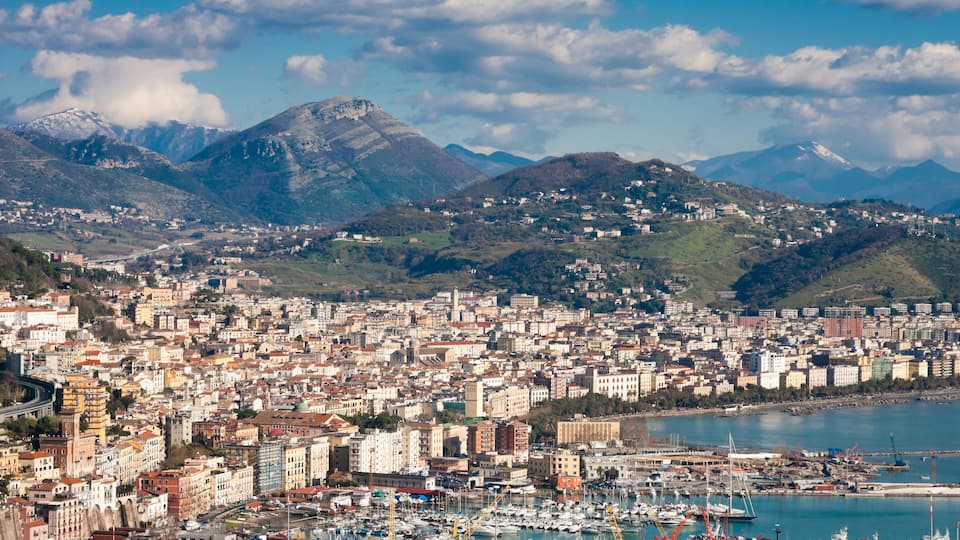A coastal city with a marina, mountains in the background, and a mix of historic and modern architecture.