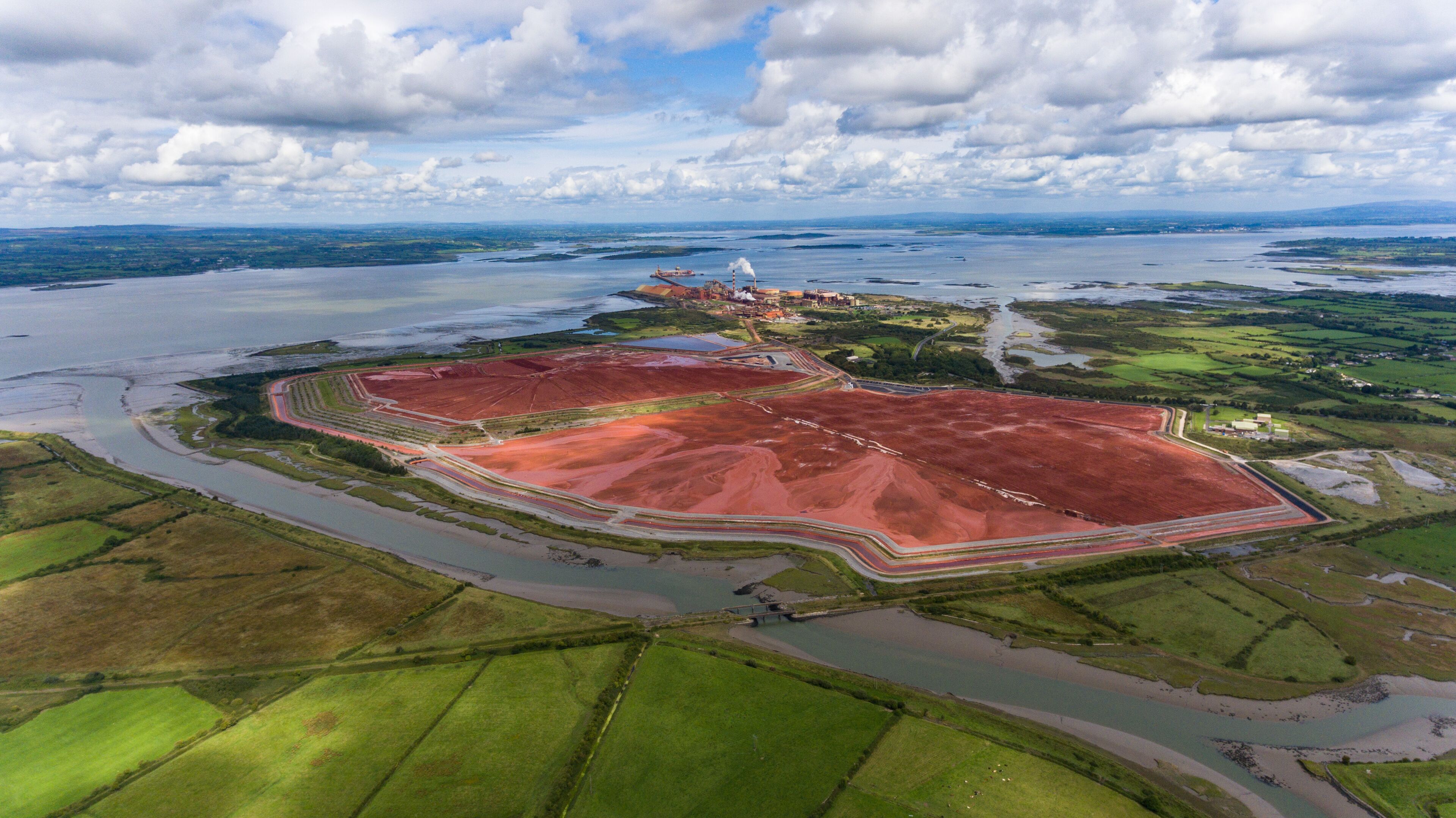 An aerial view of a coastal area with a large red mining site, a bridge, and a distant ship.