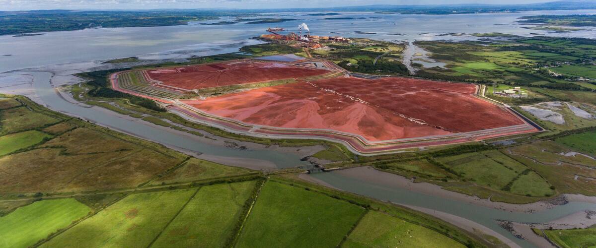 An aerial view of a coastal area with a large red mining site, a bridge, and a distant ship.