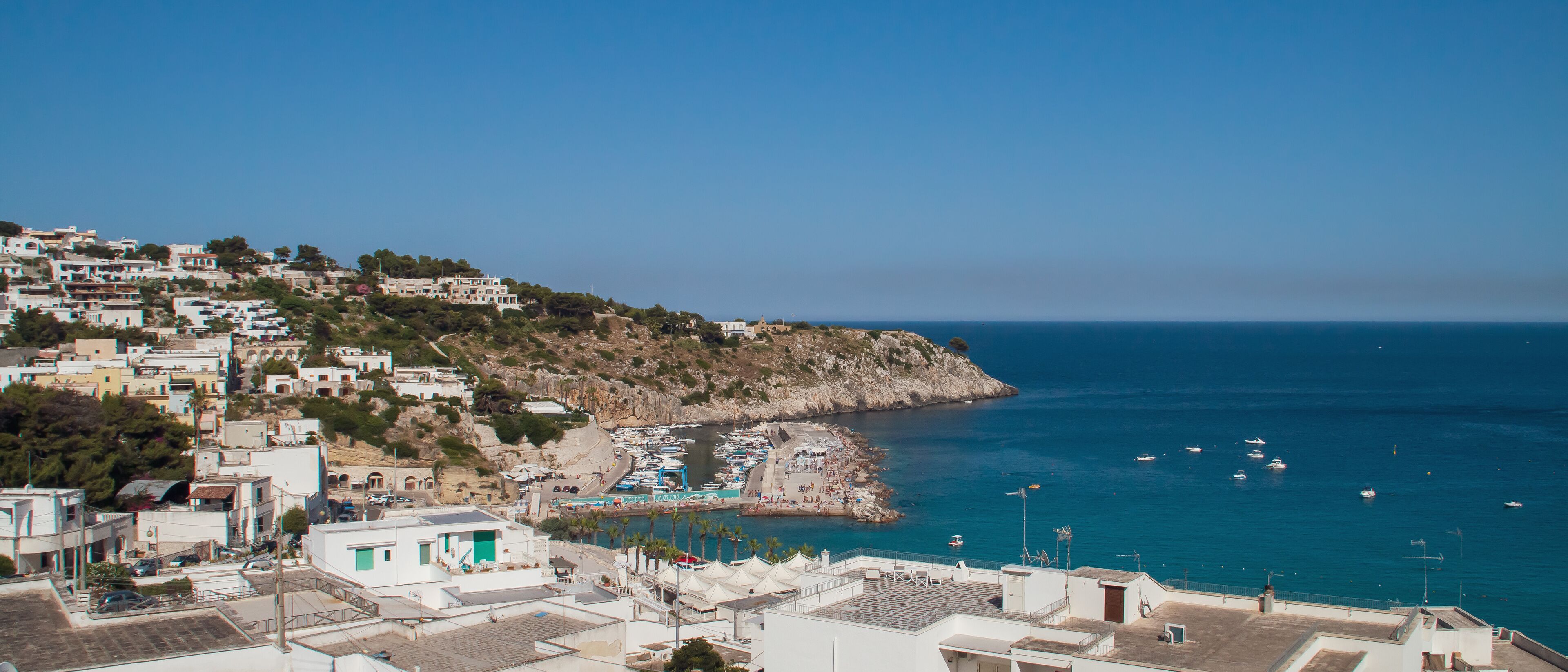 Vista del puerto deportivo de la ciudad de Castro en Lecce, Italia. Barcos atracados en el muelle y fondeados en las proximidades. Bañistas tomando el sol en el puerto.