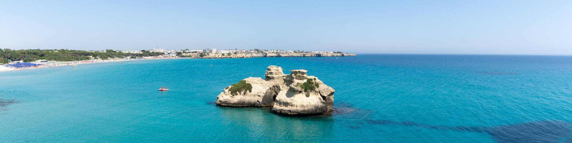 A coastal landscape with clear turquoise waters, a rocky outcrop, and a distant shoreline with buildings.