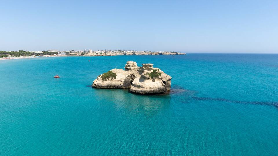 A coastal landscape with clear turquoise waters, a rocky outcrop, and a distant shoreline with buildings.