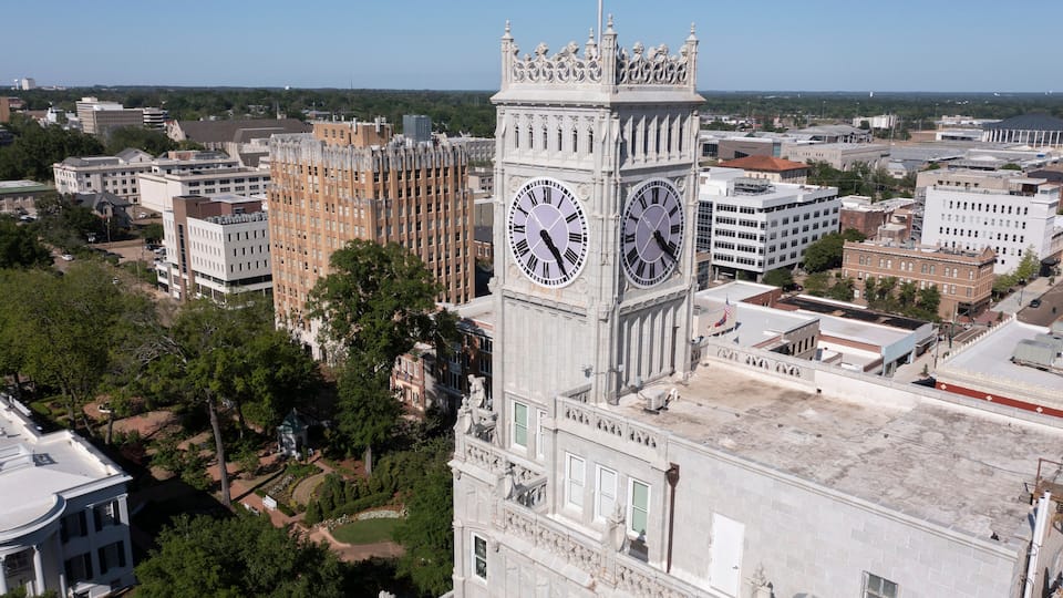 A clock tower with Roman numerals and a large clock face, surrounded by urban buildings and greenery.