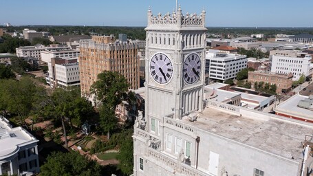 A clock tower with Roman numerals and a large clock face, surrounded by urban buildings and greenery.