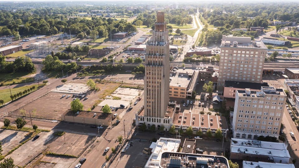 An aerial view of a cityscape with a prominent tall building with a unique design, surrounded by other buildings and a mix of urban and green spaces.