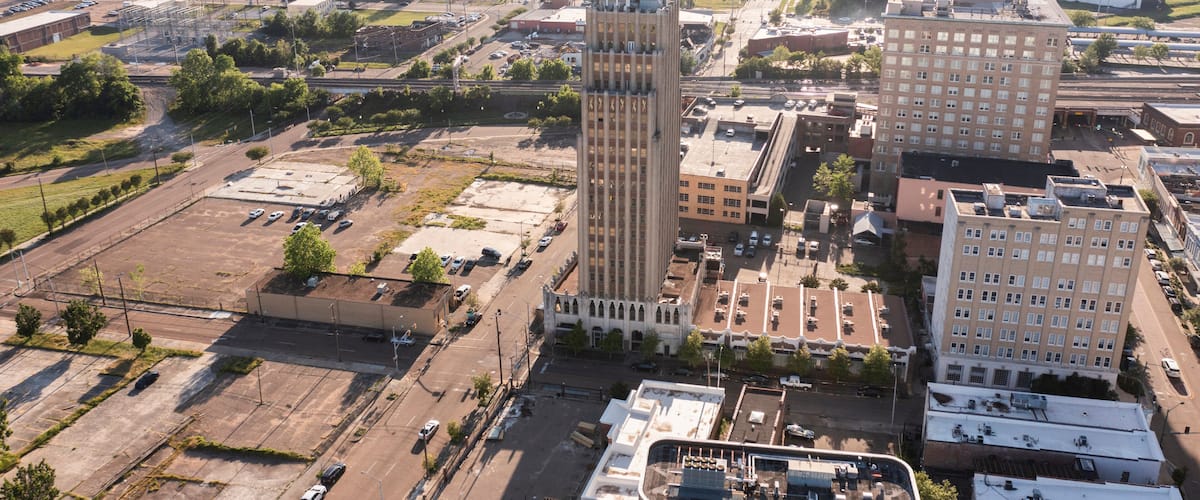 An aerial view of a cityscape with a prominent tall building with a unique design, surrounded by other buildings and a mix of urban and green spaces.