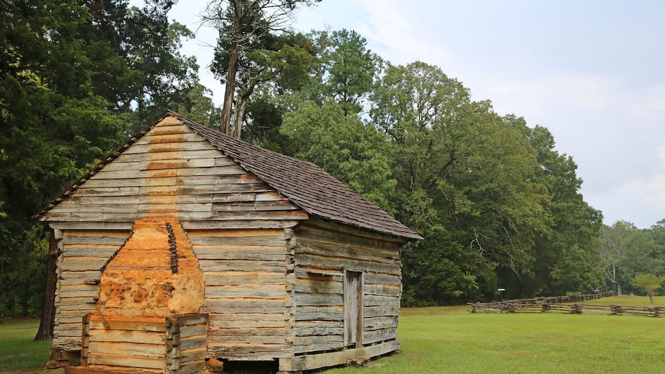 A rustic wooden cabin with a steeply pitched roof, surrounded by lush green trees and a grassy field.