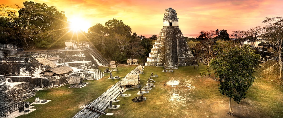 Un complejo de pirámides mayas con una pirámide central imponente, estructuras más pequeñas alrededor y un sol poniéndose al fondo.