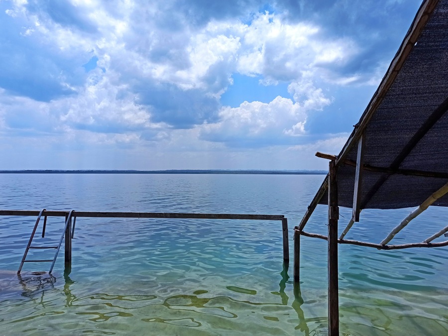 A wooden structure extending over clear blue water with a distant shoreline under a partly cloudy sky.