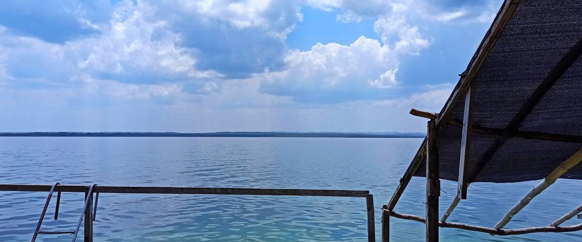 A wooden structure extending over clear blue water with a distant shoreline under a partly cloudy sky.