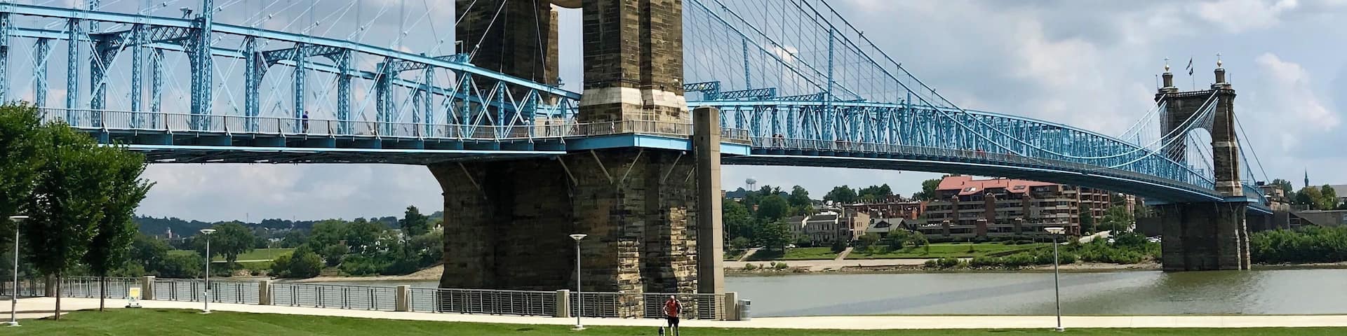 A large suspension bridge with two towers, a river below, and a grassy area in the foreground.