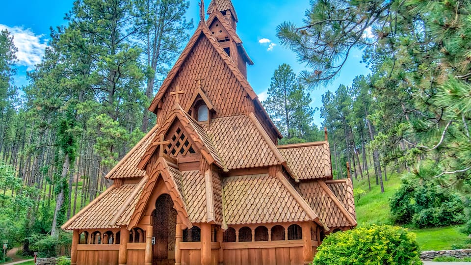 A wooden house with a steep roof and a pointed tower, surrounded by pine trees.