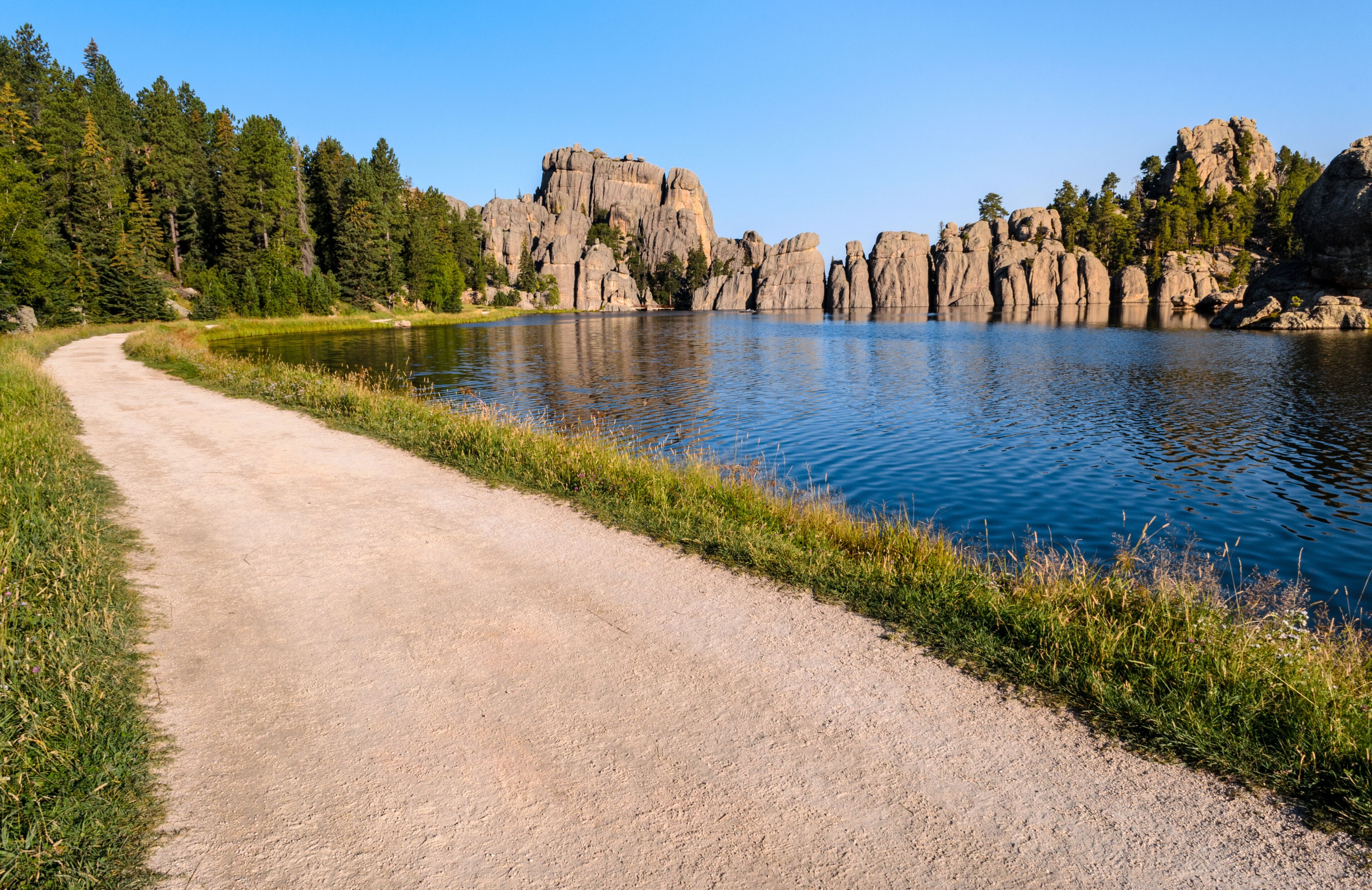 A gravel path beside a calm lake with rocky cliffs and dense forest in the background.