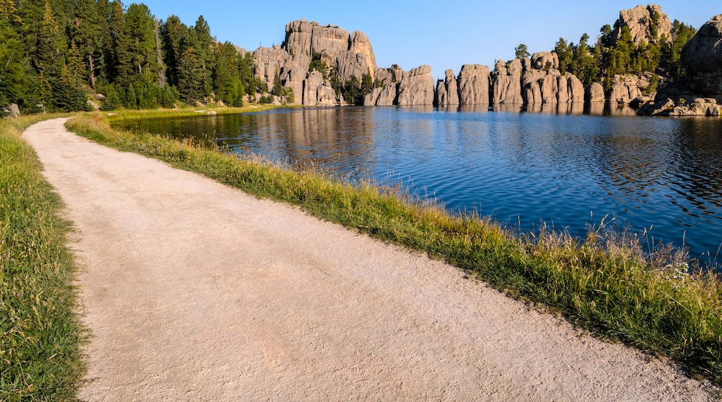 A gravel path beside a calm lake with rocky cliffs and dense forest in the background.