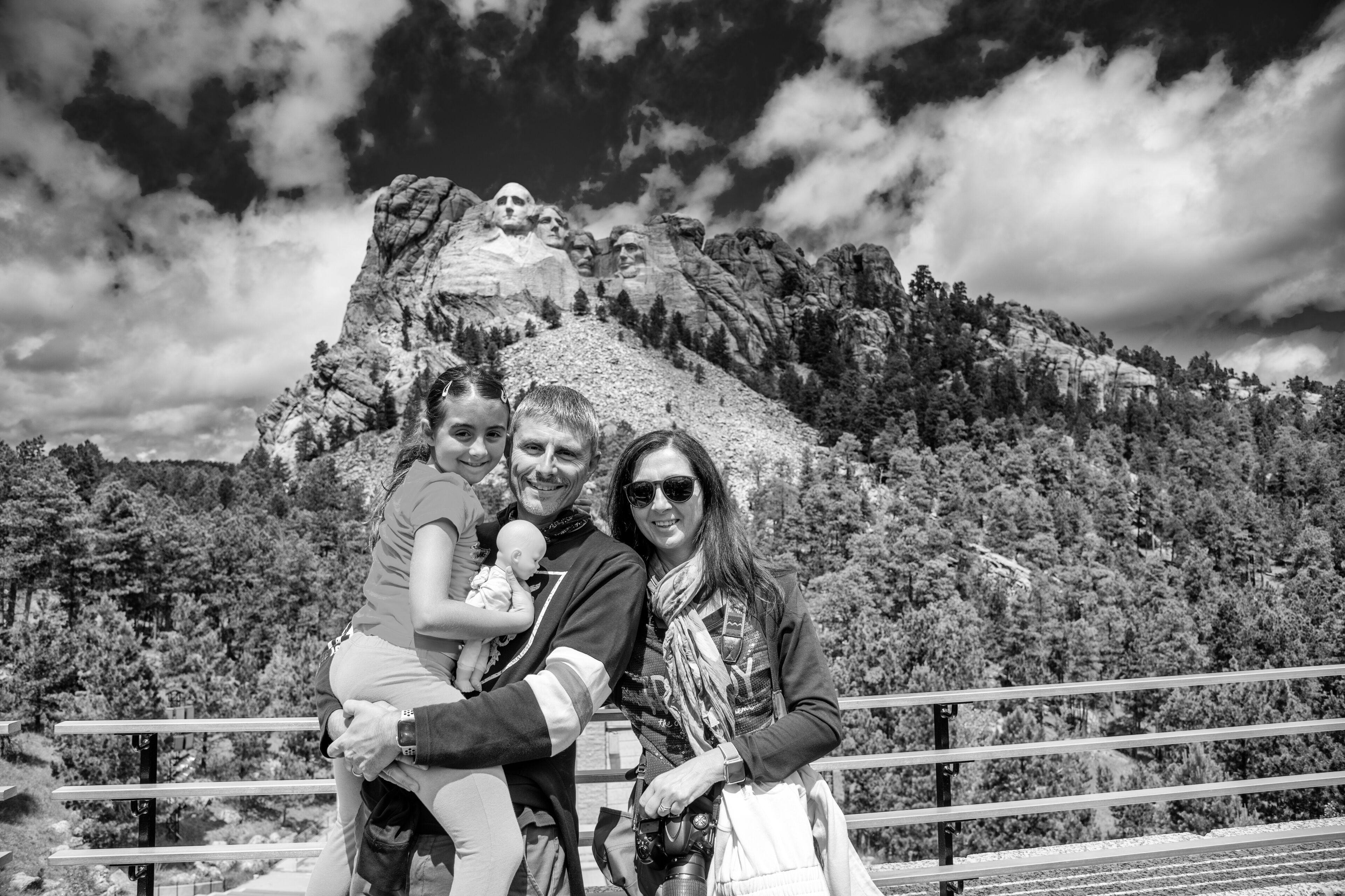 A family with a child, a man, and a woman standing in front of a mountain with a rocky peak and forested slopes.